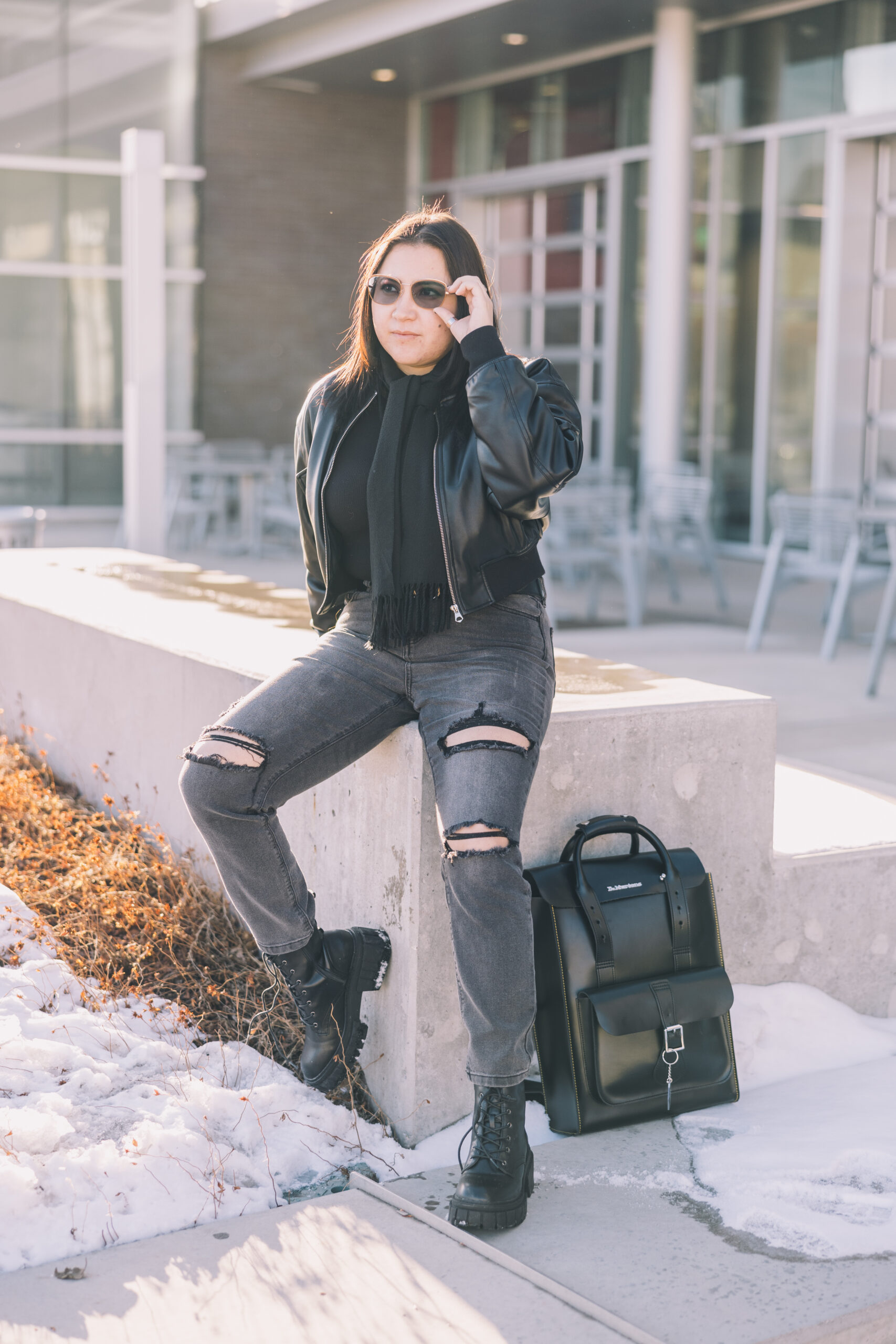 A woman leans against a concrete short wall and adjusts her sunglasses while her backpack is by her feet.