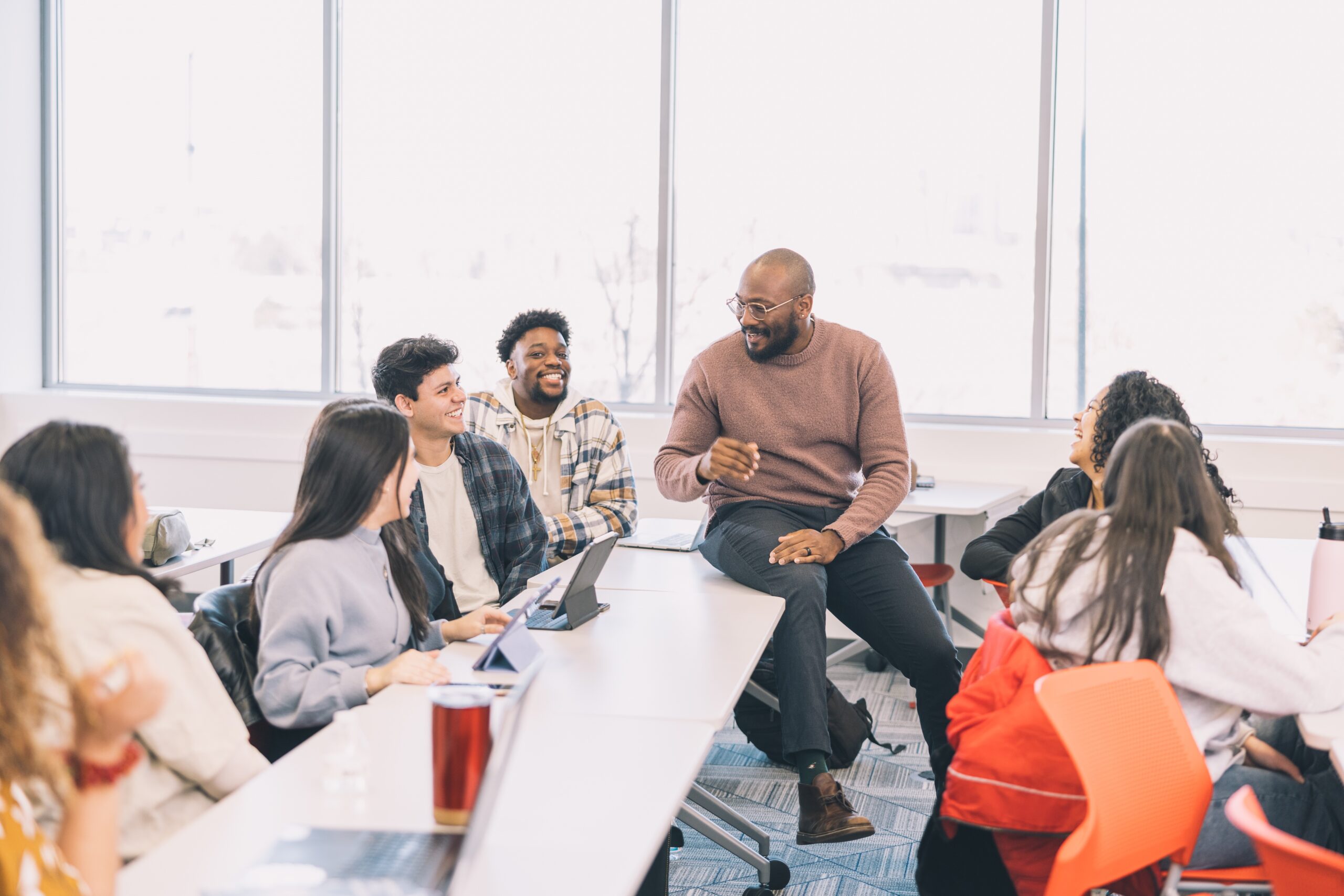 A professor sits on a table in a classroom, having a lively conversation with several students.