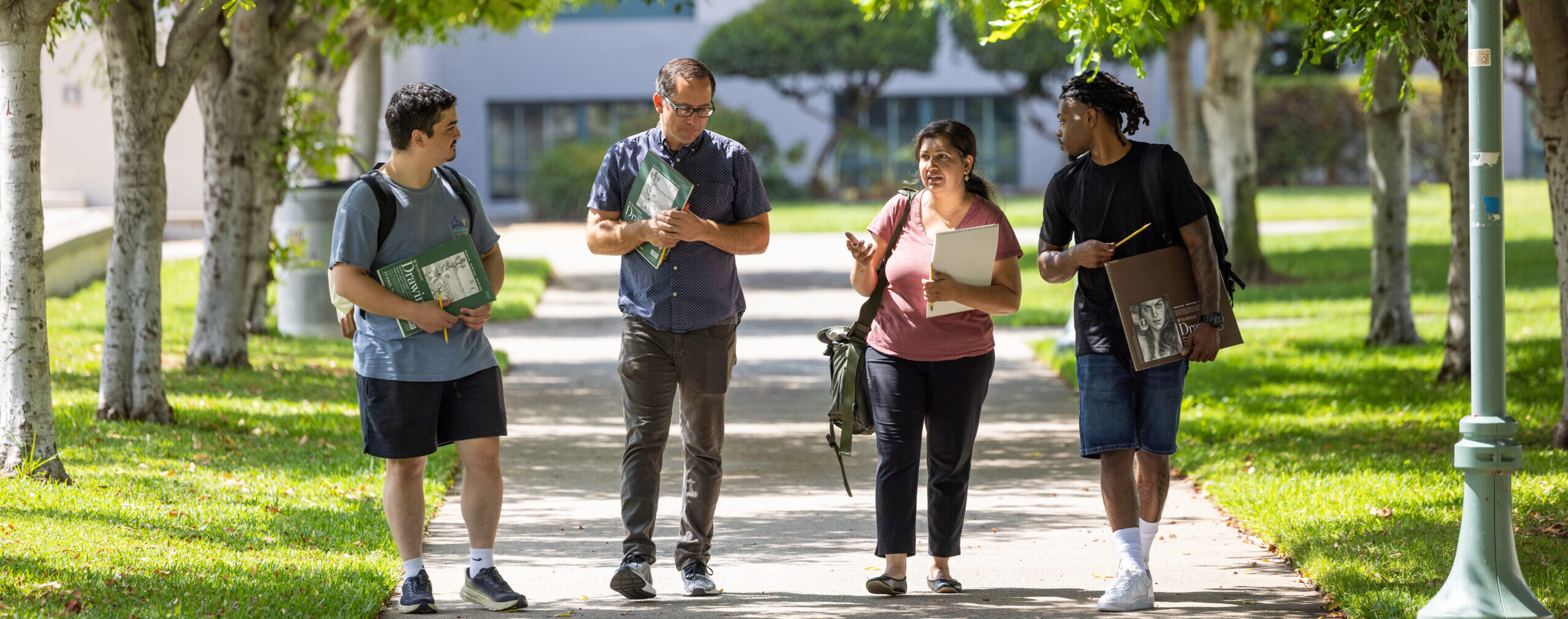 Multiethnic group of staff and students walking a path under a canopy of tree branches on a college campus.