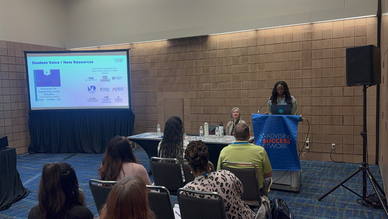 Alauna presents at a podium during an Advising Success Network session. A slide titled “Student Voice / New Resources” is projected on the screen beside her, and attendees are seated and listening. A blue ASN banner covers the front of the podium.